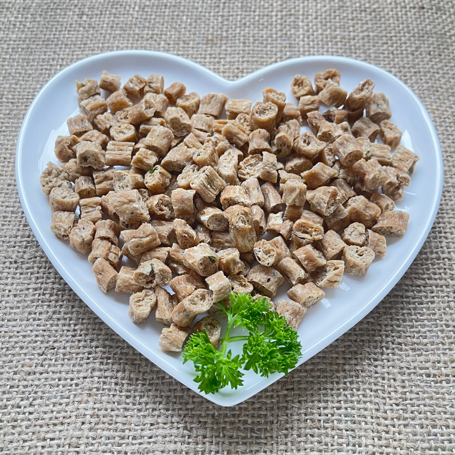 Small bite sized cat treats displayed with a sprig of parsley on a white heart shaped plate.