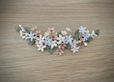 Tocado de novia de porcelana de unos 22 cm de longitud compuesto por flores blancas con toques en rosa, flores azules, flores rosa con centro burdeos y hojas verde seco.