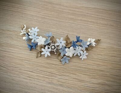 Tocado de novia de unos 20 cm de longitud compuesto por flores en tonos azules y blancos y hojas doradas alrededor de una estrella central dorada con perlas y cristales transparentes.
