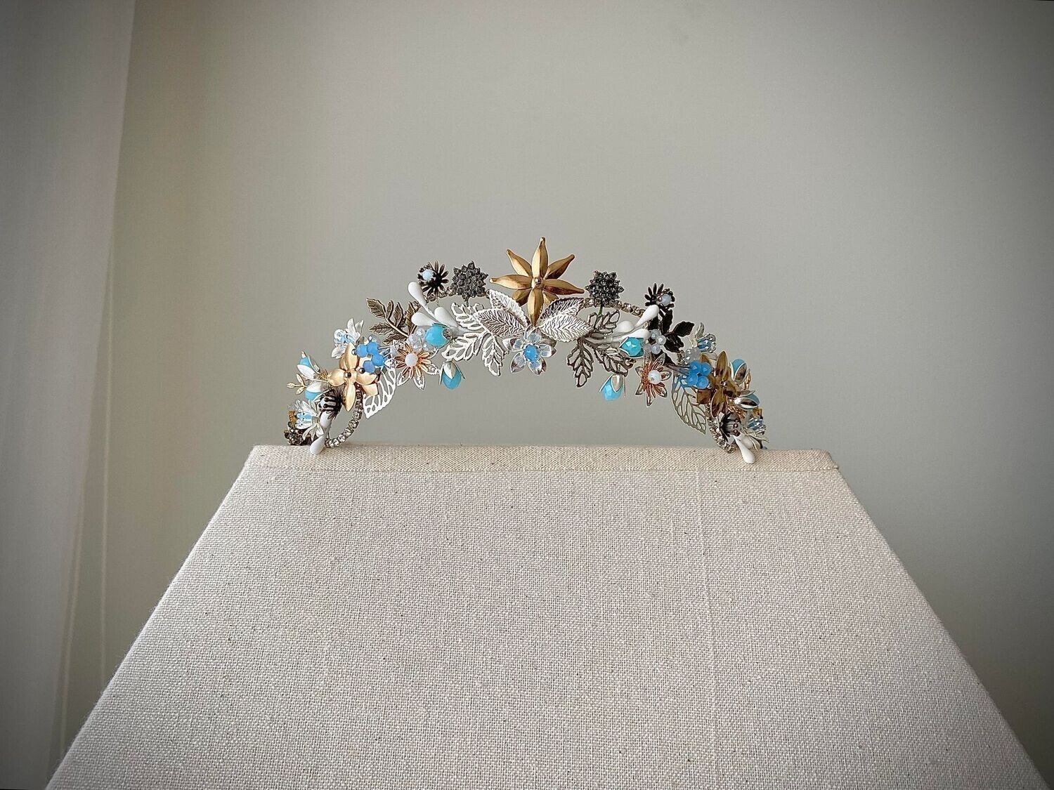 Tiara de metal y porcelana muy ligera compuesta por múltiples detalles en diferentes tonos de plata y dorado con pequeños cristales azules y matices en blanco. En el centro está adornada por una flor que puede cambiarse por una estrella u otro detalle.

Queda preciosa si la combinas con horquillas en la parte trasera de la cabeza ya que se conseguimos equilibrar el look aportando luminosidad en la zona posterior.

Su peculiaridad es que puede utilizarse también como tocado trasero por su tamaño, consiguiendo con una misma pieza lucir dos looks.