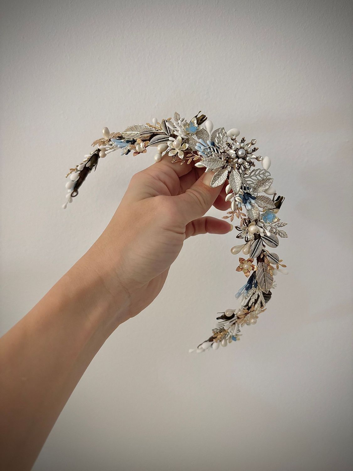 Tiara de novia de metal y porcelana compuesta por diferentes detalles dentro de los que destacan las flores, las hojas y los pistilos con una estrella central. Este tocado de novia se caracteriza por ser una tiara de boda muy tupida con un ligero acabado en pico en el centro.