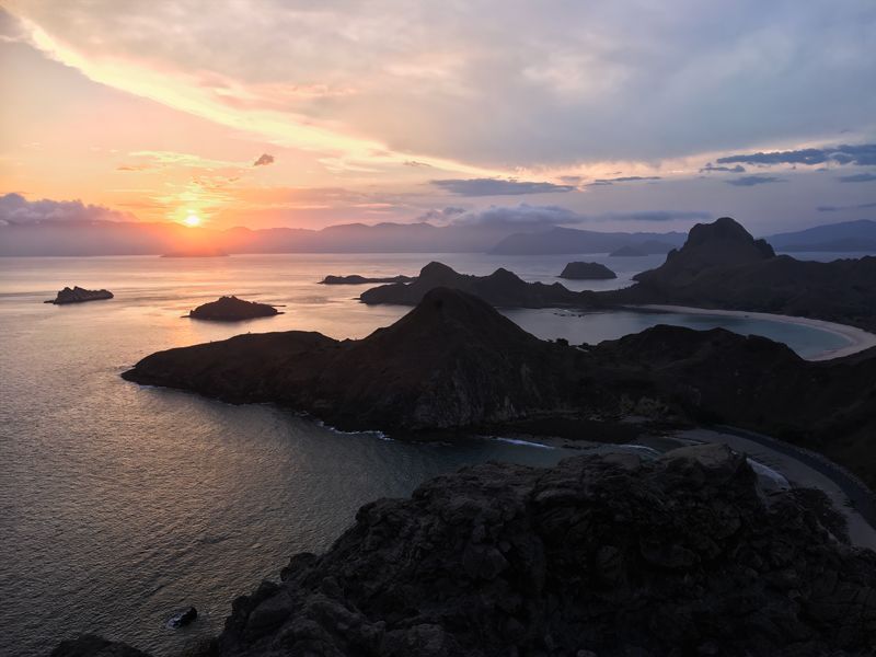 Komodo National Park - Sunset over Pulau Padar The sun is setting near the horizon, casting a warm glow across the sky and reflecting on the water. Several dark, rugged islands and hills are silhouetted against the colorful sky, with calm waters surrounding them.