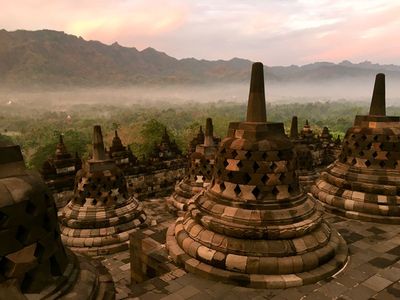 Borobudur Temple is a marvel of ancient architecture. The image captures several stupas on the upper terraces, set against a backdrop of lush greenery and distant mountains under a colorful sky at sunrise.