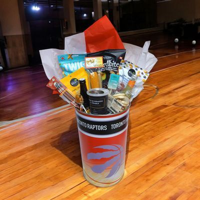 Raptor basketball gift basket in red team bucket on wooden floor with snacks, mirror and wall in the background
