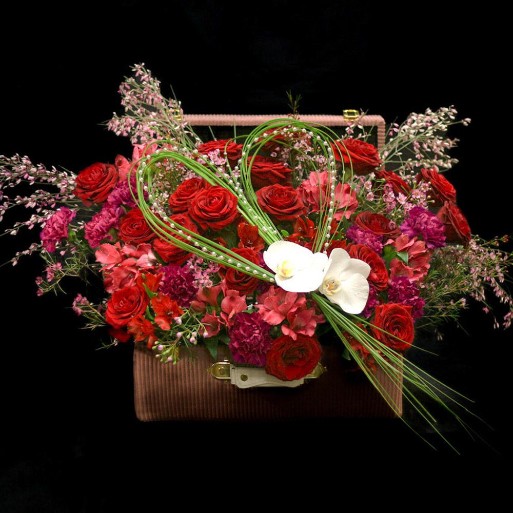 Un somptueux arrangement de fleurs de saison dans les tons rouges et fuchsia, présenté dans une valise en velours raffinée.