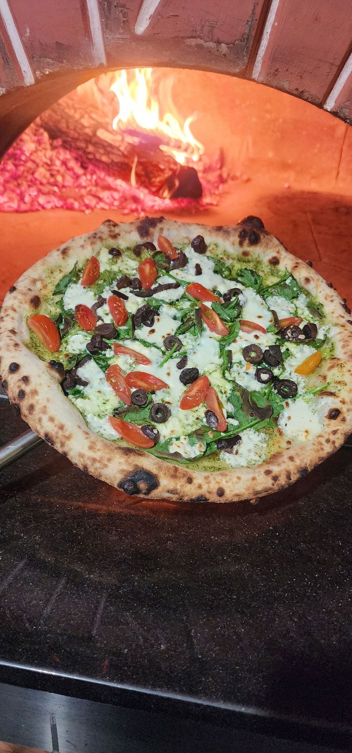 Neapolitan style Pizza with Pesto, Black Olives, Tomatoes, Spinach pictured in front of a wood fired oven