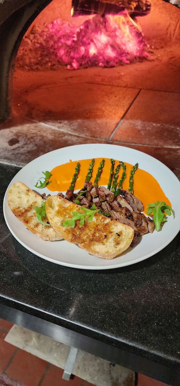 Plate with steak cuts, bread on a bed of asparagus and creamy hot sauce held in front of a wood fired oven