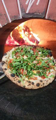 Neapolitan style pizza with calabrese salami and arugula held in front of a wood burning oven 