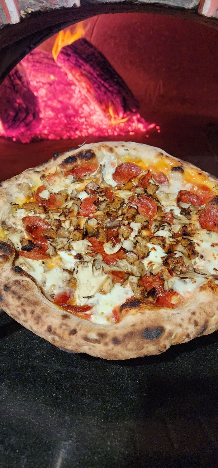 Neapolitan Style Pizza with pepperoni, sausage, and mushrooms held in front of a wood fired oven 