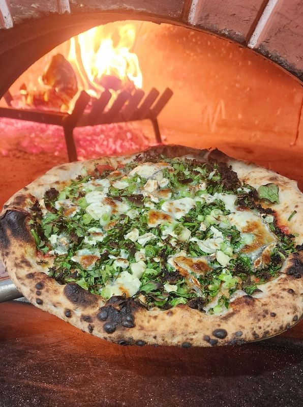 Neapolitan Style Pizza with spinach, kale, humboldt fog goat cheese and san marzano held in front of a wood burning oven 