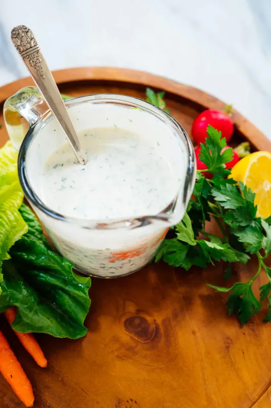 ranch sauce in a glass jar with a stirrer on a board surrounded by herbs