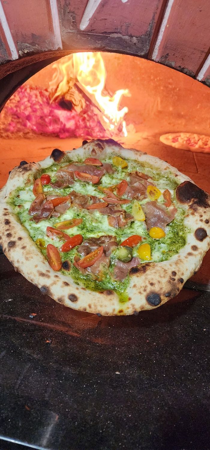 Pesto e Prosciutto Neapolitan style pizza with pesto, fresh mozzarella, prosciutto and tomatoes held in front of a wood fired oven