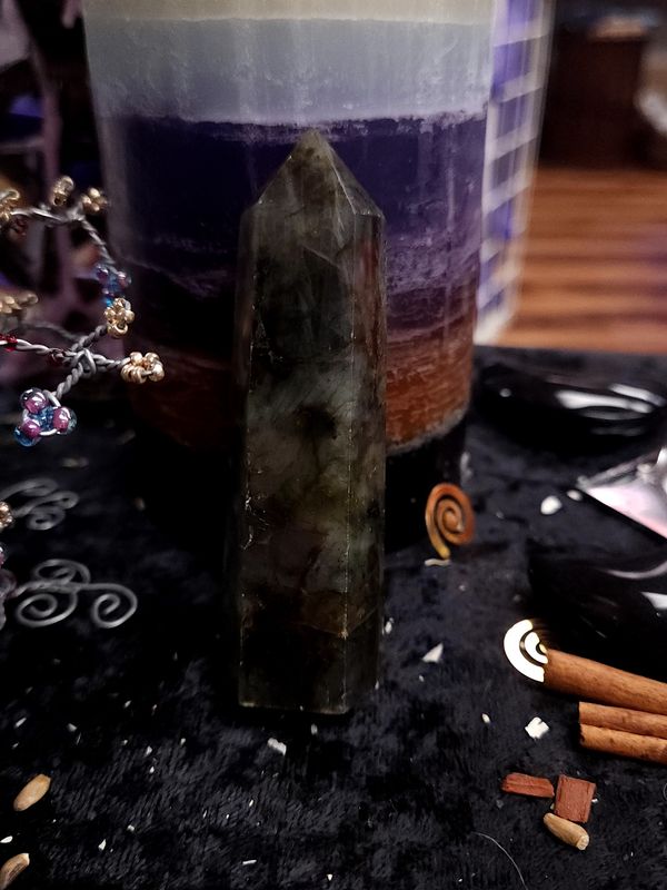 Twilight Veil Labradorite Tower 88mm Dark green-gray 88mm labradorite tower standing on a black altar cloth before a layered ritual candle.