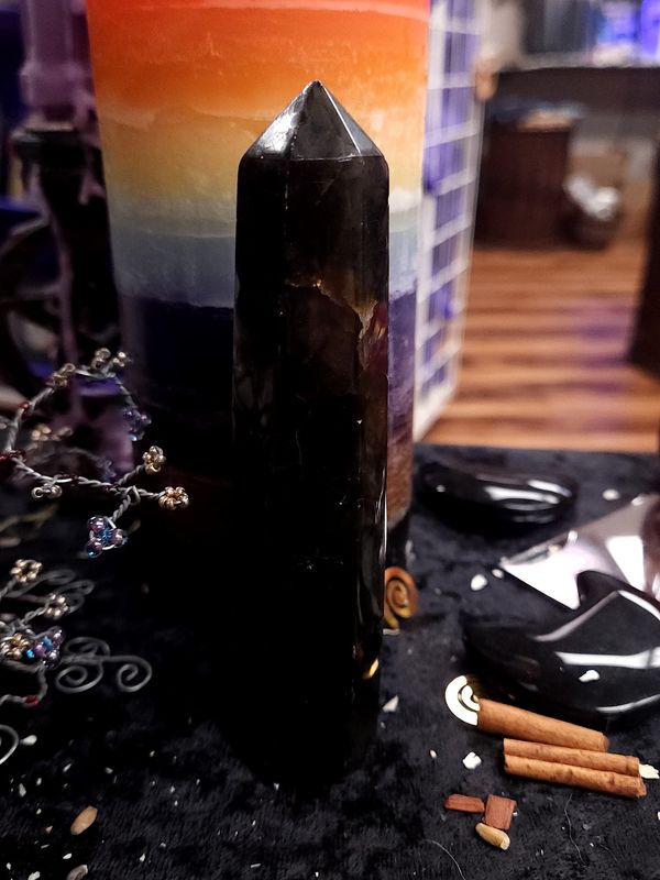 Shadow Ward Labradorite Tower 132mm Tall, very dark 132mm labradorite tower on a black altar cloth in front of a multicolored pillar candle.