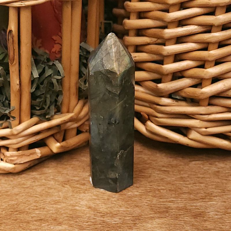 Dawn Ember Labradorite Tower 68mm Small 68mm labradorite tower with soft flash at the tip, standing on a wooden surface in front of woven wicker baskets.