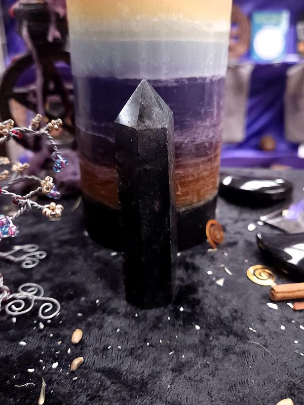 Starlit Shield Astrophyllite Tower 93mm 93mm dark astrophyllite tower with a smoky, sparkling tip on a black altar cloth before a rainbow pillar candle.