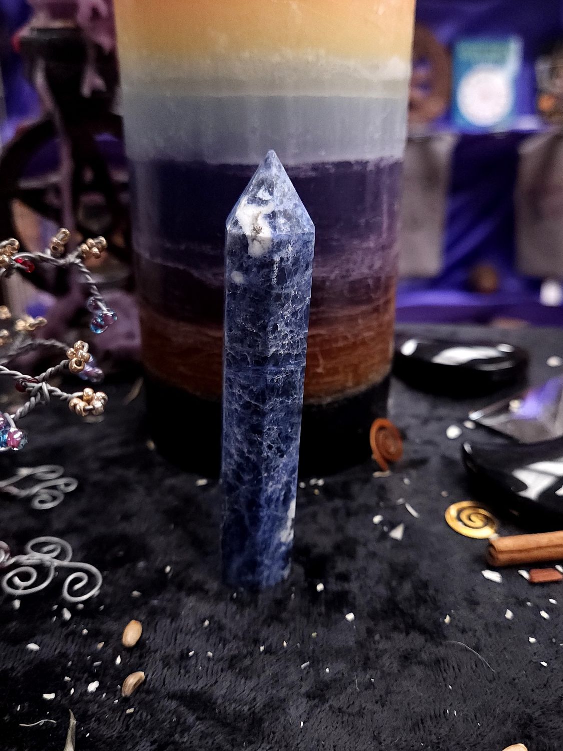 Starpoint Sodalite Tower 94mm 94mm blue sodalite tower with a white-tipped point on a black altar cloth in front of a rainbow pillar candle.