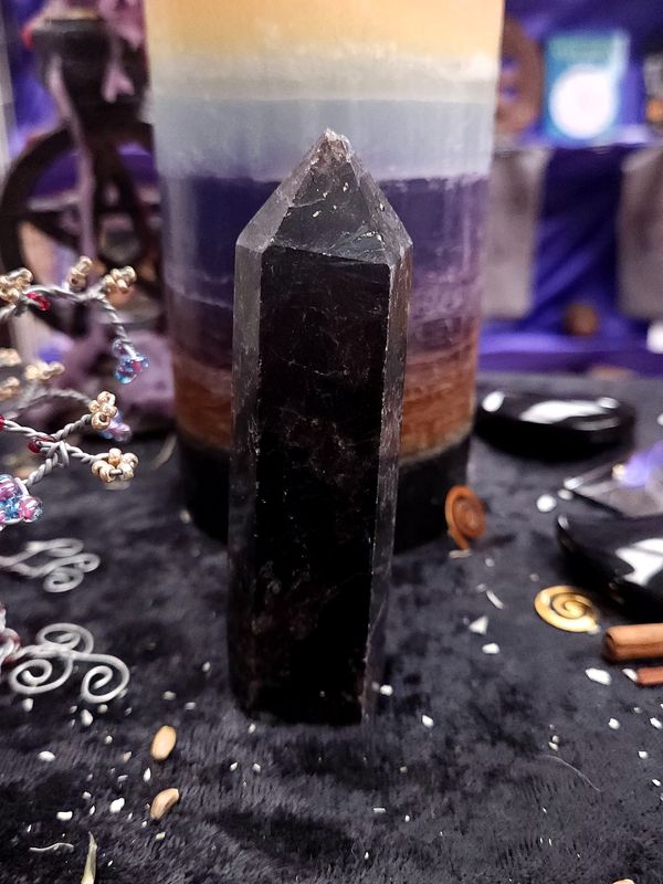 Cosmic Gate Astrophyllite Tower 100mm Tall 100mm dark astrophyllite tower with a slightly sparkling tip on a black altar cloth before a rainbow pillar candle.
