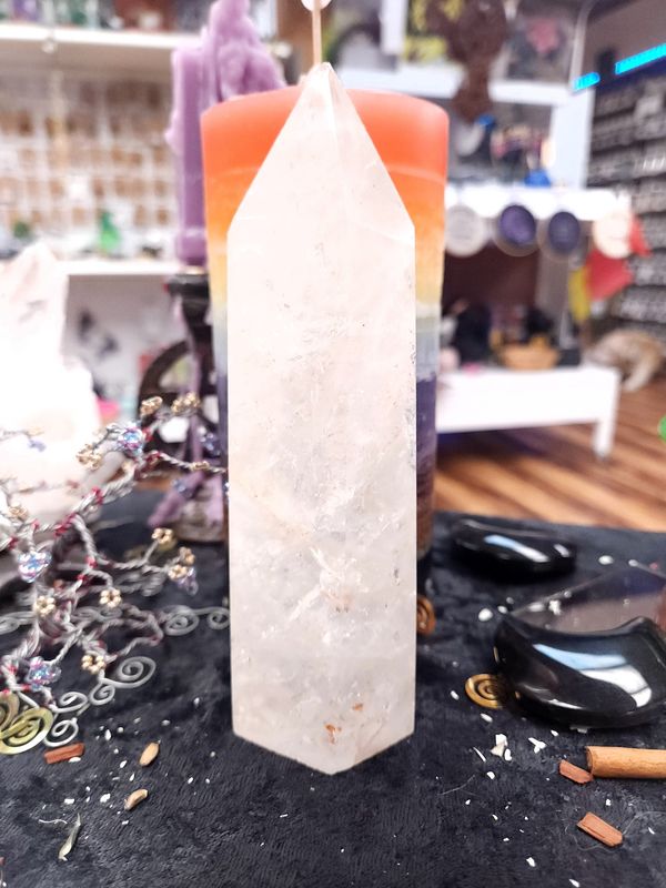 Tall Clear Quartz Tower 165mm Tall clear quartz tower on a black altar cloth in front of a rainbow pillar candle, with crystal trees and shop shelves