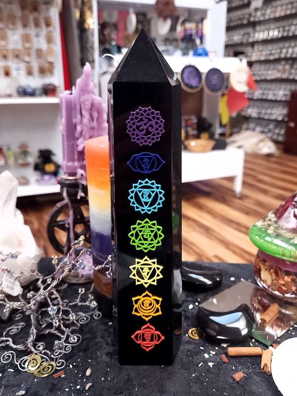 Chakra Obsidian Tower Tall black obsidian tower etched with rainbow chakra symbols standing on a black altar cloth in a crystal shop, with candles