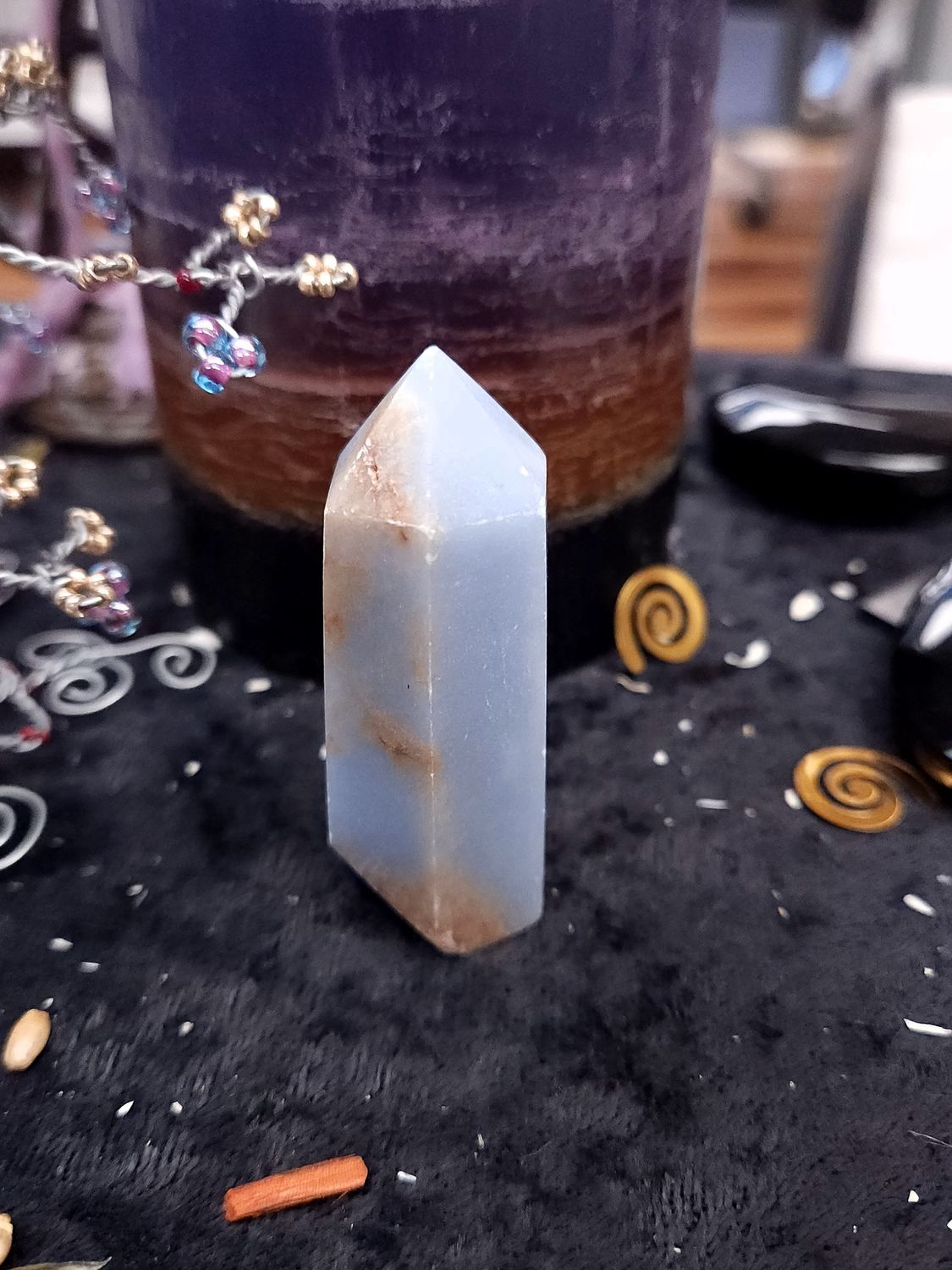 Angelite Crystal Tower 62mm Soft blue angelite crystal tower with brown streaks on a black altar cloth in front of a purple layered candle and decor