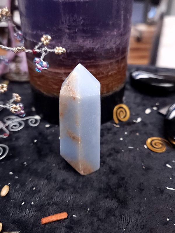 Angelite Crystal Tower 62mm Soft blue angelite crystal tower with brown streaks on a black altar cloth in front of a purple layered candle and decor