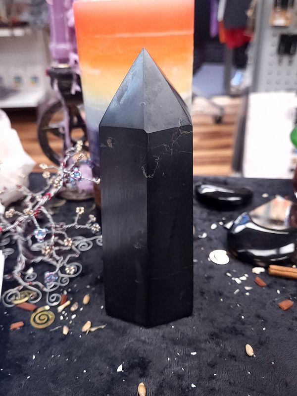 Shungite Crystal Tower 142mm Tall black shungite crystal tower on a black altar cloth in front of a rainbow pillar candle with crystal tree and decor
