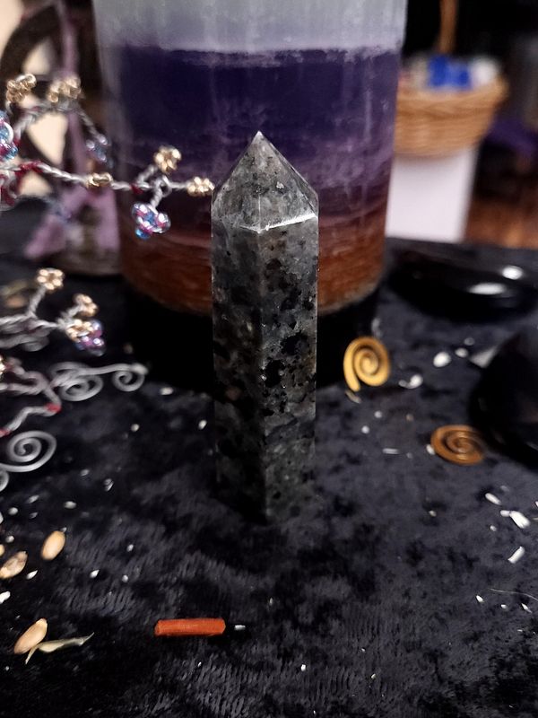 Yooperlite Tower 80mm Tall grey and black yooperlite crystal tower on a black altar cloth in front of a purple layered candle with wire decor