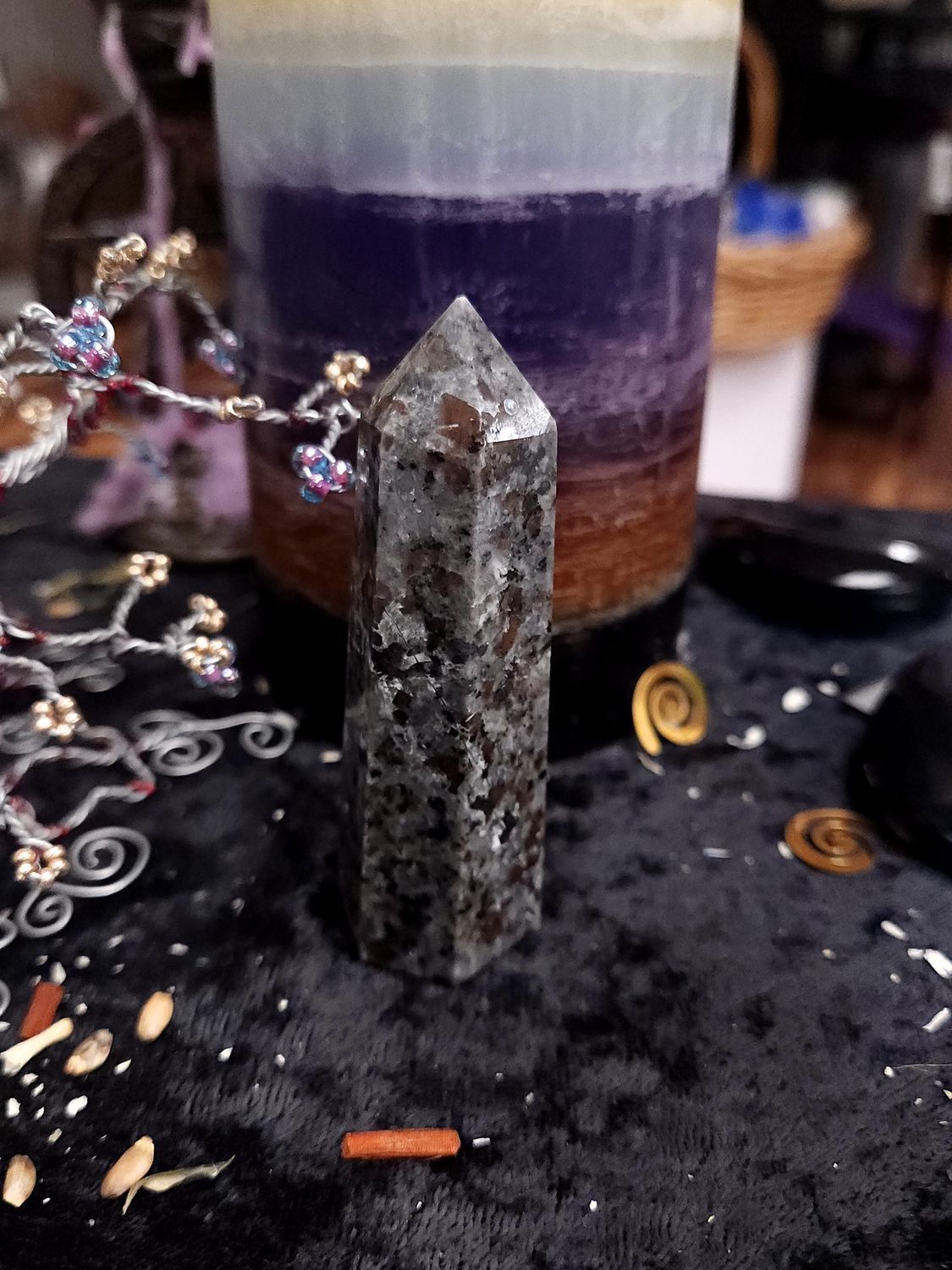 Grey and black yooperlite crystal tower on a black altar cloth in front of a purple layered candle with wire gemstone tree 