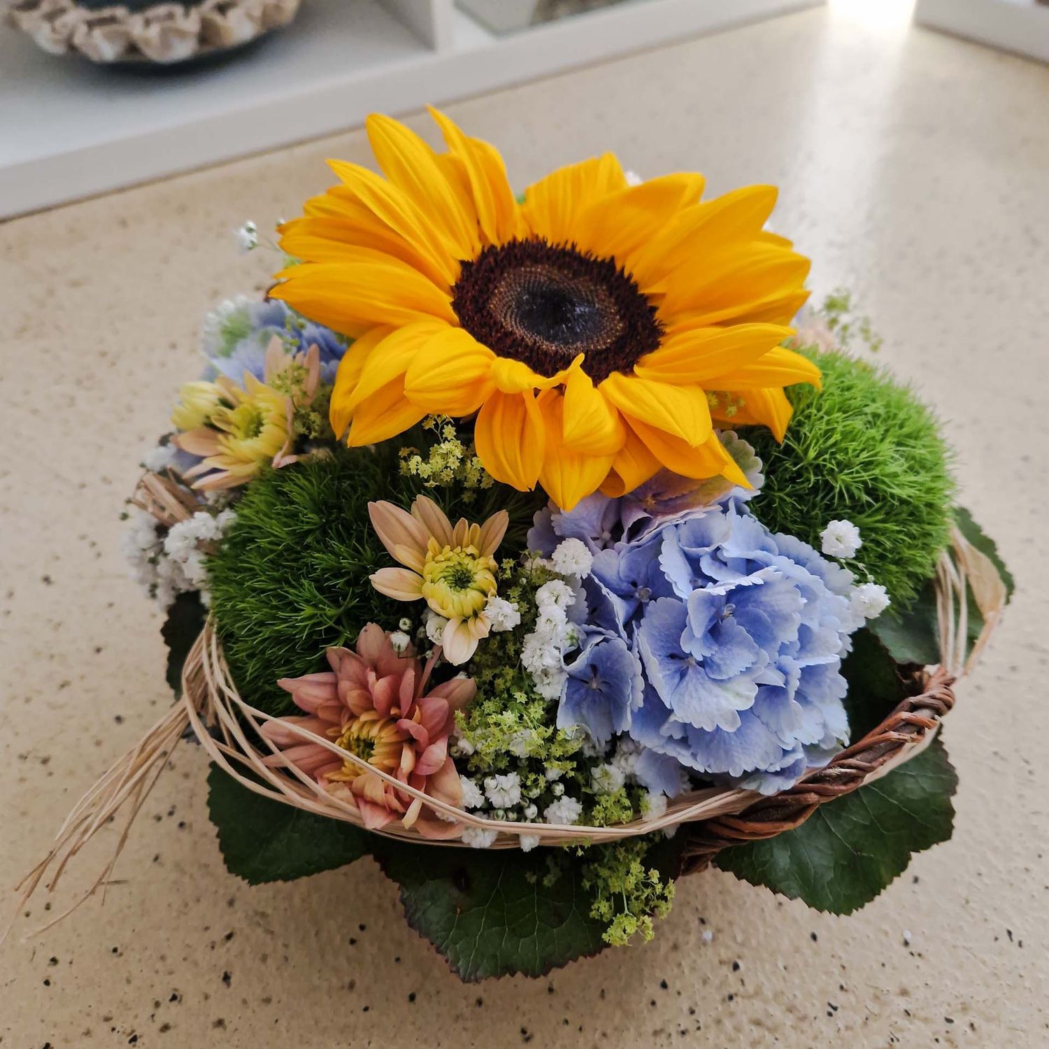 Gesteck mit Sonnenblume, in Korb