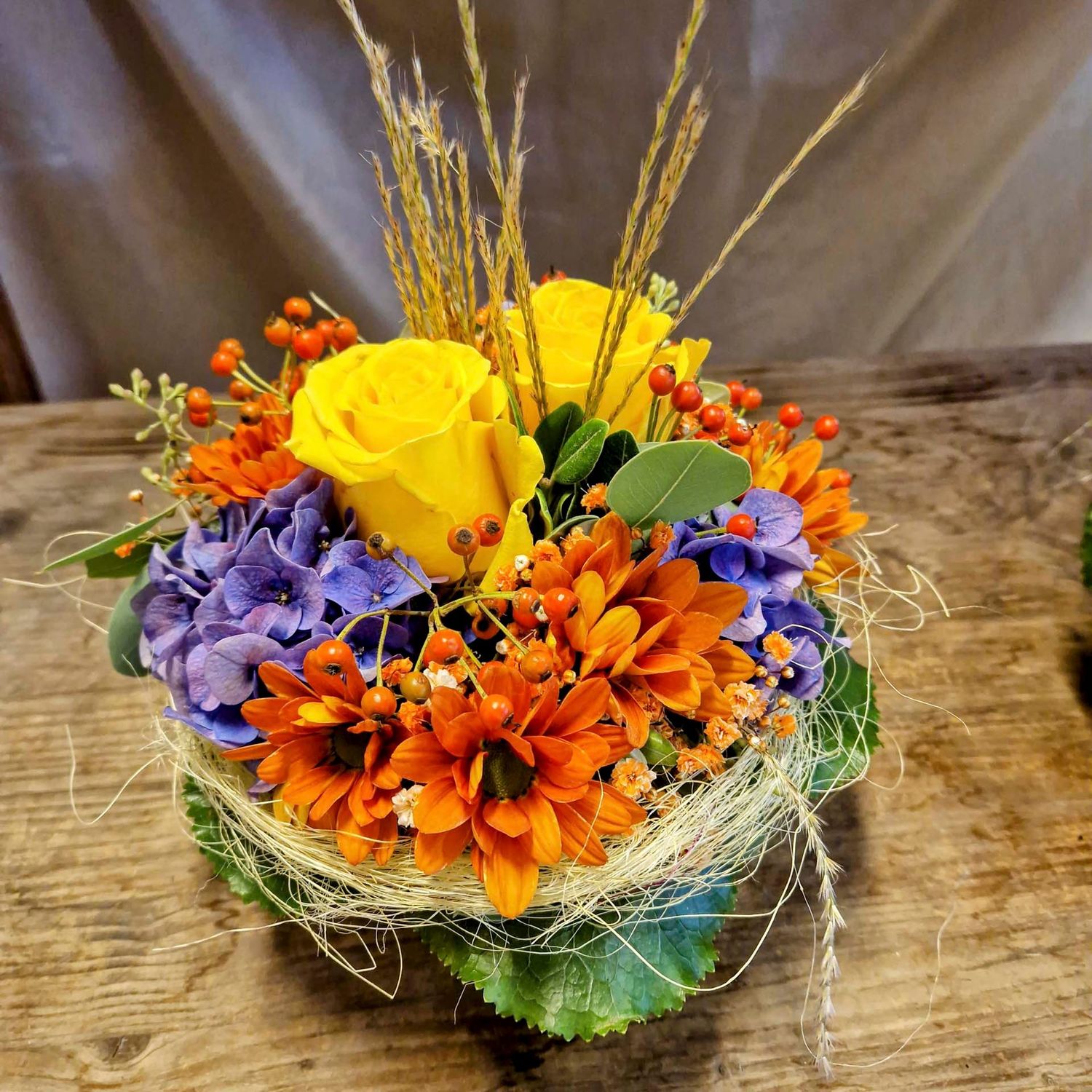 Gesteck mit zwei Rosen und gemischten Blumen, Beeren und Halmen, gelb/orange/blau, in Korb (Øca.22cm) Gesteck mit zwei Rosen und gemischten Blumen, Beeren und Halmen, gelb/orange/blau, in Korb (Øca.22cm)