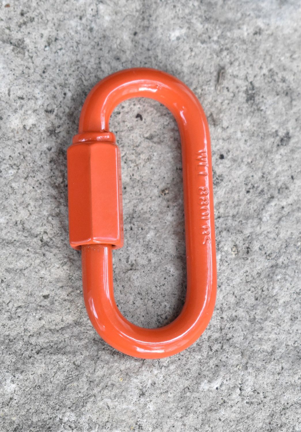 Close-up of a 1/4 inch quick link metal connector with threaded screw lock, resting on a concrete background.