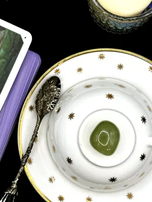 Translucent olive green healerite tumbled stone on a white teacup and saucer with gold stars.