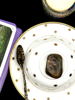 Black moonstone tumbled stone in dark gray-brown with a silvery sheen on a white teacup and saucer with gold stars.