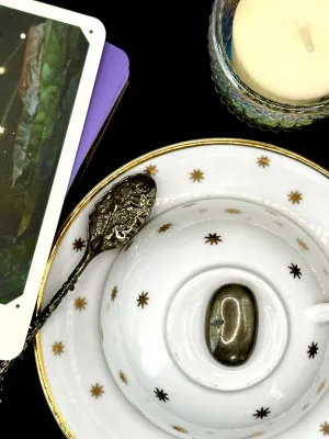 Brassy metallic pyrite tumbled stone with a polished oval shape on a white teacup and saucer with gold stars.