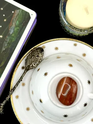 Red sardonyx tumbled stone in deep orange-red with faint white banding on a white teacup and saucer with gold stars.