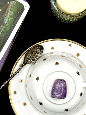 Purple lepidolite tumbled stone with white flecks on a white teacup and saucer with gold stars.