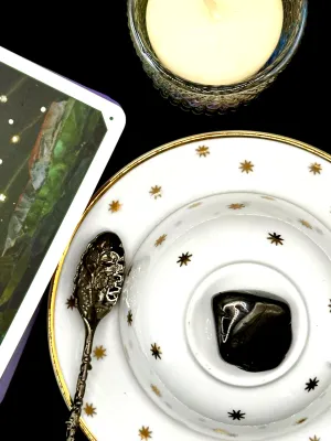 Glossy black jet tumbled stone with angled faces on a white teacup and saucer with gold stars.