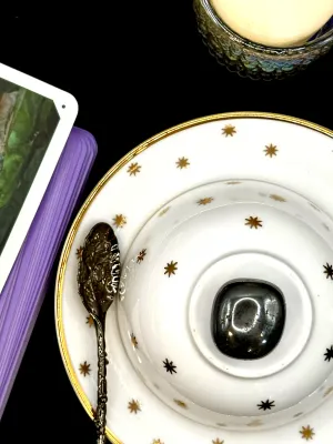 Metallic dark gray hematite tumbled stone with a mirror-like polish on a white teacup and saucer with gold stars.