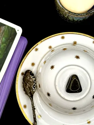 Black golden sheen obsidian tumbled stone with a soft gold flash on a white teacup and saucer with gold stars.