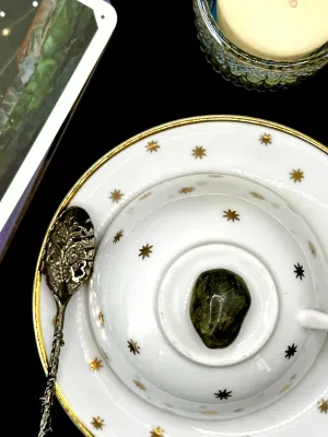 Dark mossy green nephrite jade tumbled stone with mottled spots on a white teacup and saucer with gold stars.
