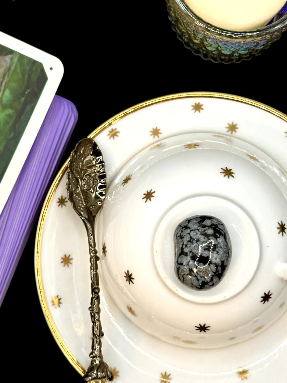 Black snowflake obsidian tumbled stone with gray snowflake spots on a white teacup and saucer with gold stars.