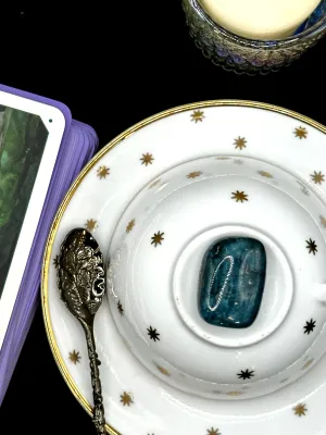 Deep teal blue apatite tumbled stone with lighter streaks on a white teacup and saucer with gold stars.