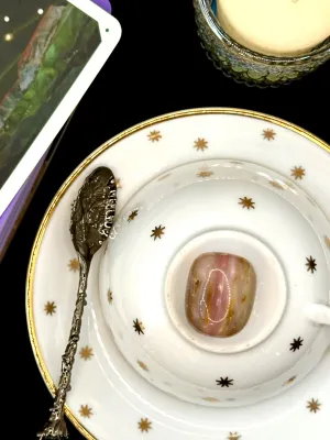 Pink opal tumbled stone banded in pink, cream, and brown on a white teacup and saucer with gold stars.