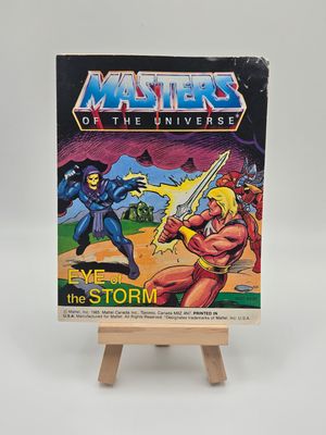Version anglaise de la couverture de la mini bande dessinée vintage &#39;Les Maîtres de l&#39;Univers&#39; intitulée &#39;Eye of the storm&#39; exposée sur un petit chevalet en bois. L&#39;illustration de la couverture montre Skeletor et Musclor (He-Man) en train de se battre, avec un arrière-plan dramatique orageux. 