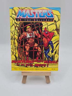 Front cover of the English version of the vintage &#39;Masters of the Universe&#39; mini comic titled &#39;Mantenna and the Menace of the Evil Horde!&#39; displayed on a small wooden easel. The cover art features He-Man caught in a net and Mantenna standing beside him.
