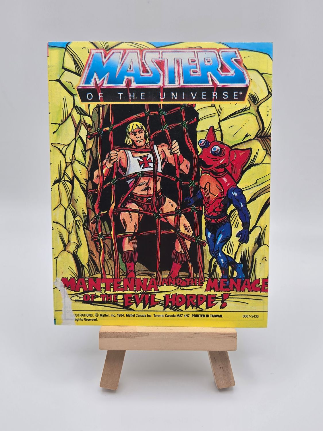Front cover of the English version of the vintage &#39;Masters of the Universe&#39; mini comic titled &#39;Mantenna and the Menace of the Evil Horde!&#39; displayed on a small wooden easel. The cover features He-Man trapped in a net with Mantenna standing beside him. Some wear is visible on the bottom left corner.