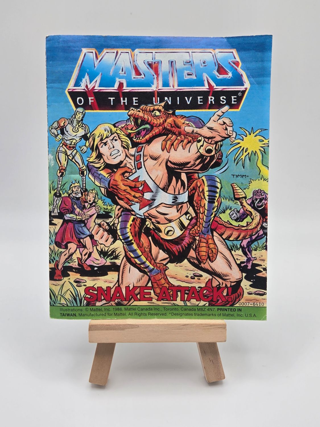 Front cover of the vintage &#39;Masters of the Universe&#39; mini comic titled &#39;Snake Attack!&#39; displayed on a small wooden easel. The cover shows He-Man fighting Rattlor, with other characters in the background.
