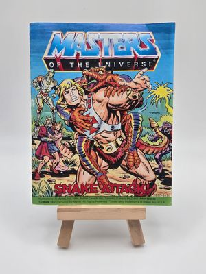 Front cover of the vintage &#39;Masters of the Universe&#39; mini comic titled &#39;Snake Attack!&#39; displayed on a small wooden easel. The cover shows He-Man fighting Rattlor, with other characters in the background.