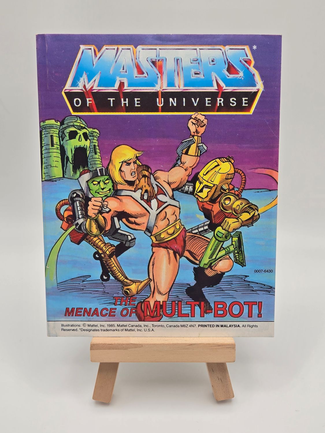 Front cover of the vintage &#39;Masters of the Universe&#39; mini comic titled &#39;The Menace of Multi-Bot!&#39; displayed on a small wooden easel. The cover features He-Man battling the robotic Multi-Bot, with Castle Grayskull in the background.